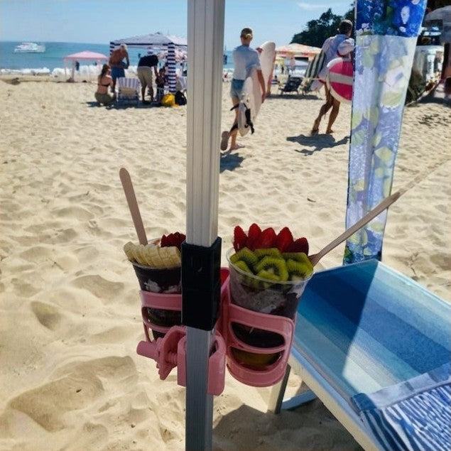 A pink beach umbrella cup holder mounted on a beach cabana pole at a sandy beach, holding two cups of iced Oakberry acai bowels with fruit slices, with beachgoers and umbrellas in the background.