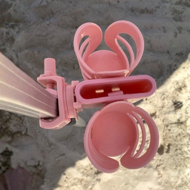 A close-up of a pink plastic 3-in-1 beach cup holder accessory attached to a beach cabana pole, shown outdoors on sandy ground with natural sunlight and shadows.