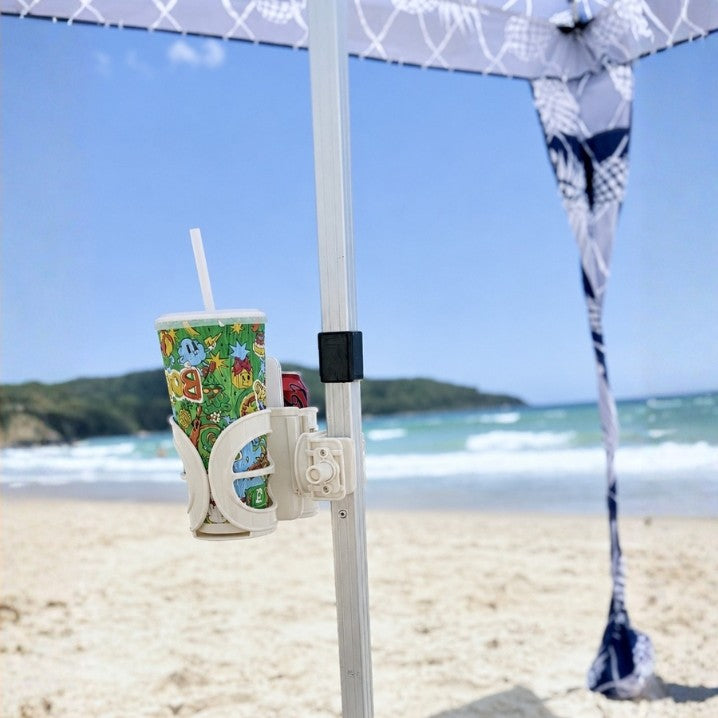 A white beach umbrella cup holder mounted on a white umbrella pole at a beach, holding a Boost Juice drink cup with a straw and can of coke with iphone in the middle.