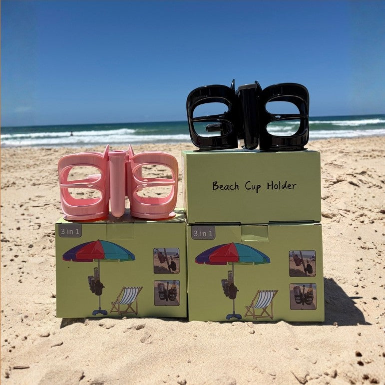 3-in-1 Beach Cup Holder 1 pink, 1 black displayed on top of their product packaging boxes on a sandy beach. The boxes are light green with illustrations of beach umbrellas and chairs, and the text 'Beach Cup Holder' is printed on the top box.