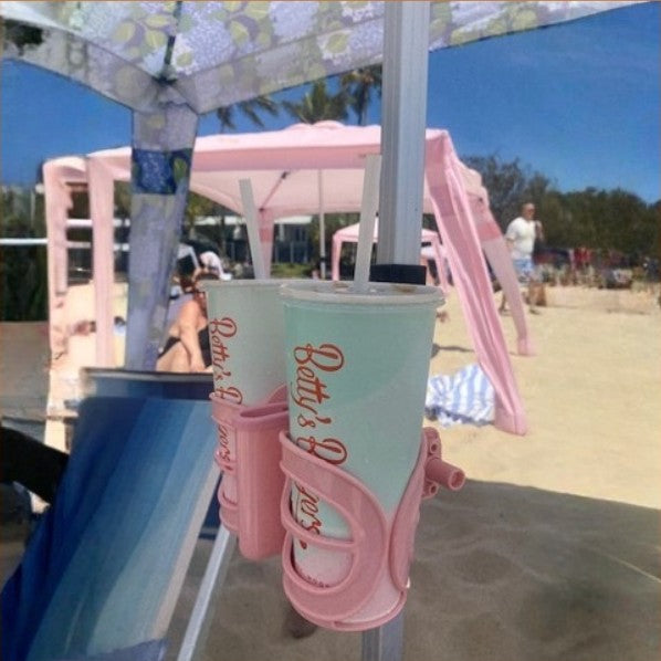Two drinks pink and mint green beach cups with straws, held in a pink holder on a beach umbrella at the beach. The cups feature the text 'Betsy's Burgers' in red script.
